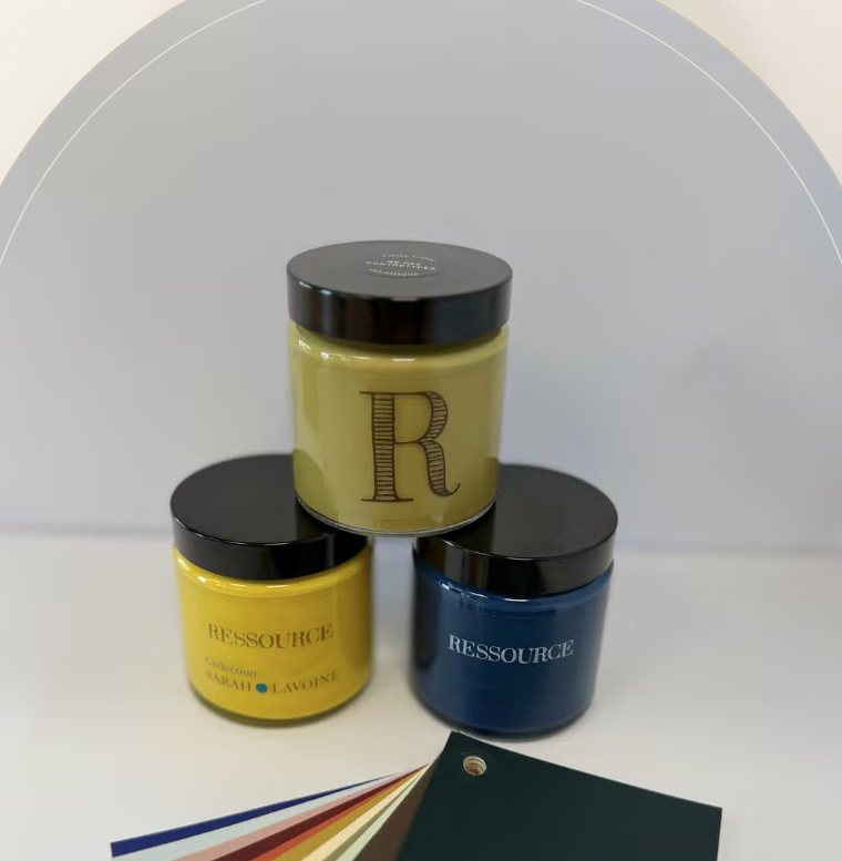 Trois pots de peinture Ressource empilés, jaune moutarde, jaune et bleu marine, devant un éventail de nuanciers